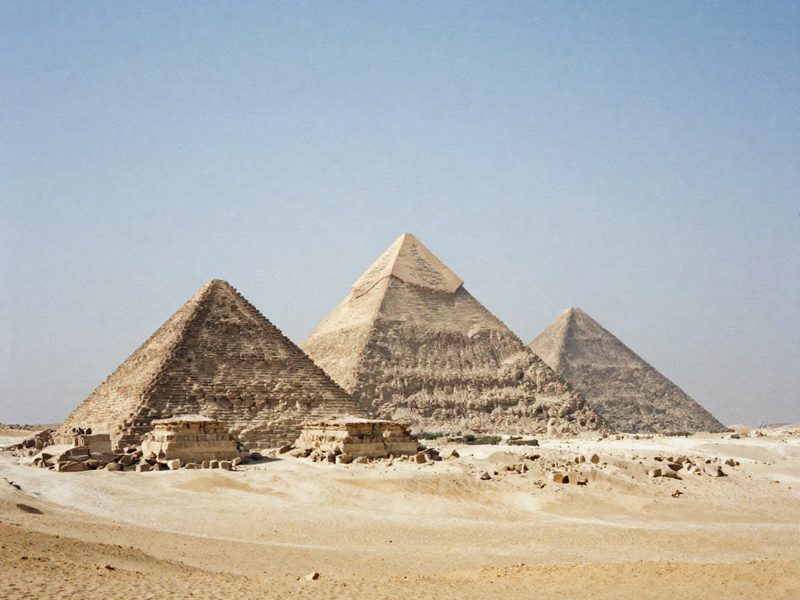 Tre piramidi di pietra allineate su un altopiano desertico al sole basso, di dimensioni decrescenti, con la sabbia increspata dal vento in primo piano