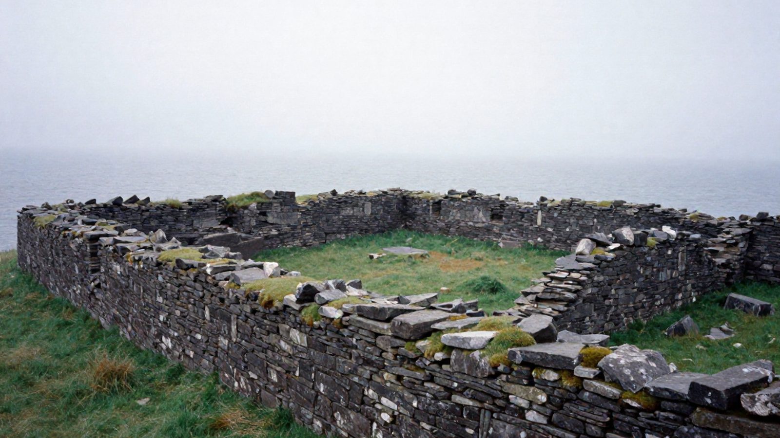 I muri in rovina di una fortezza altomedievale su un promontorio erboso battuto dal vento, sopra un mare grigio, nella nebbia